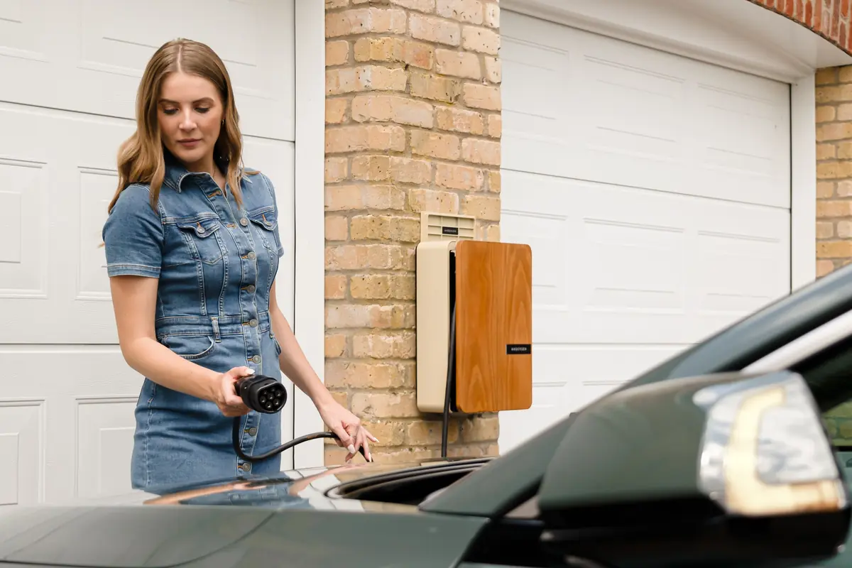 A woman holds a charging connector beside a wall charger outside her garage