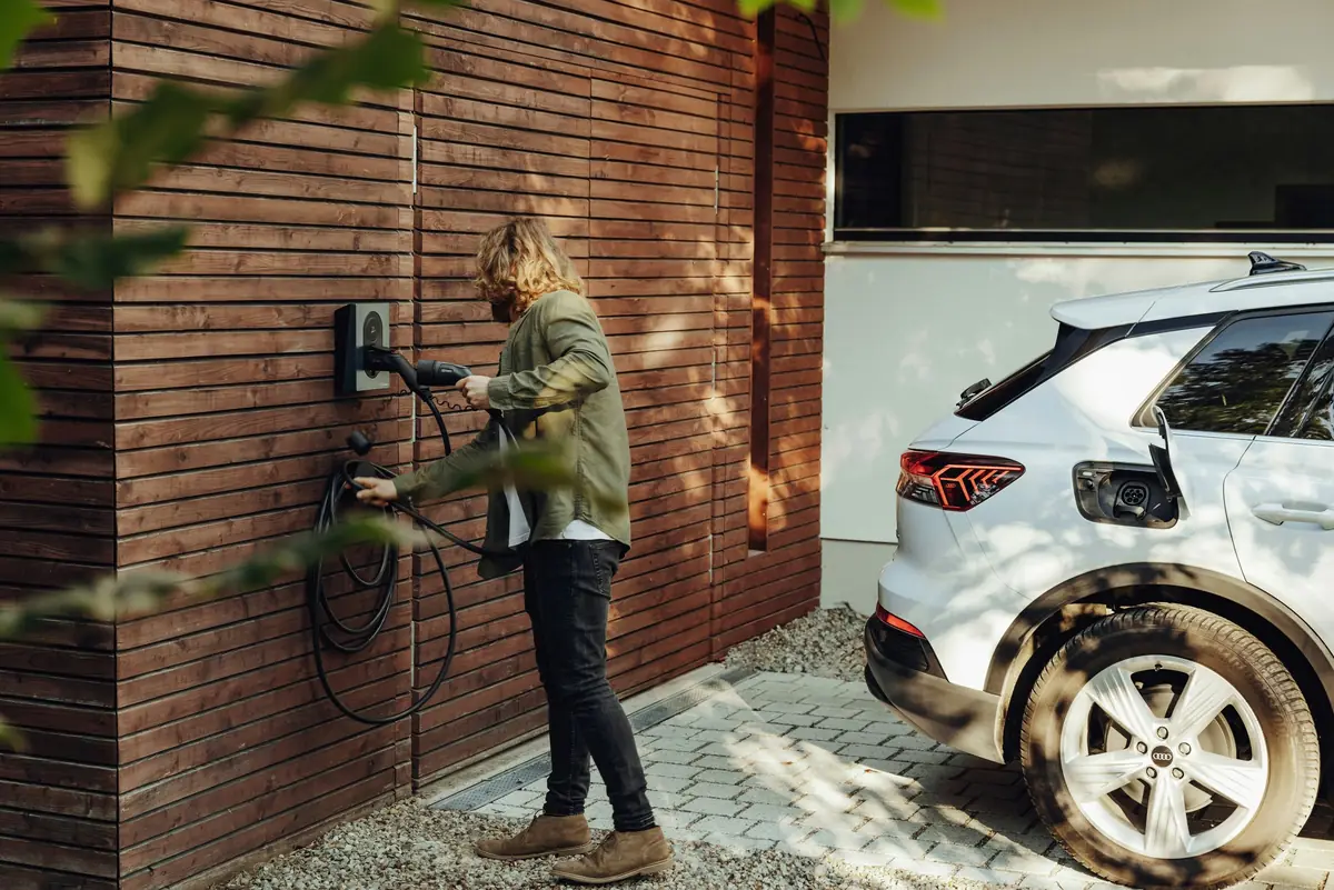 A woman plugs in an SUV at a wall charger on a timber-clad house