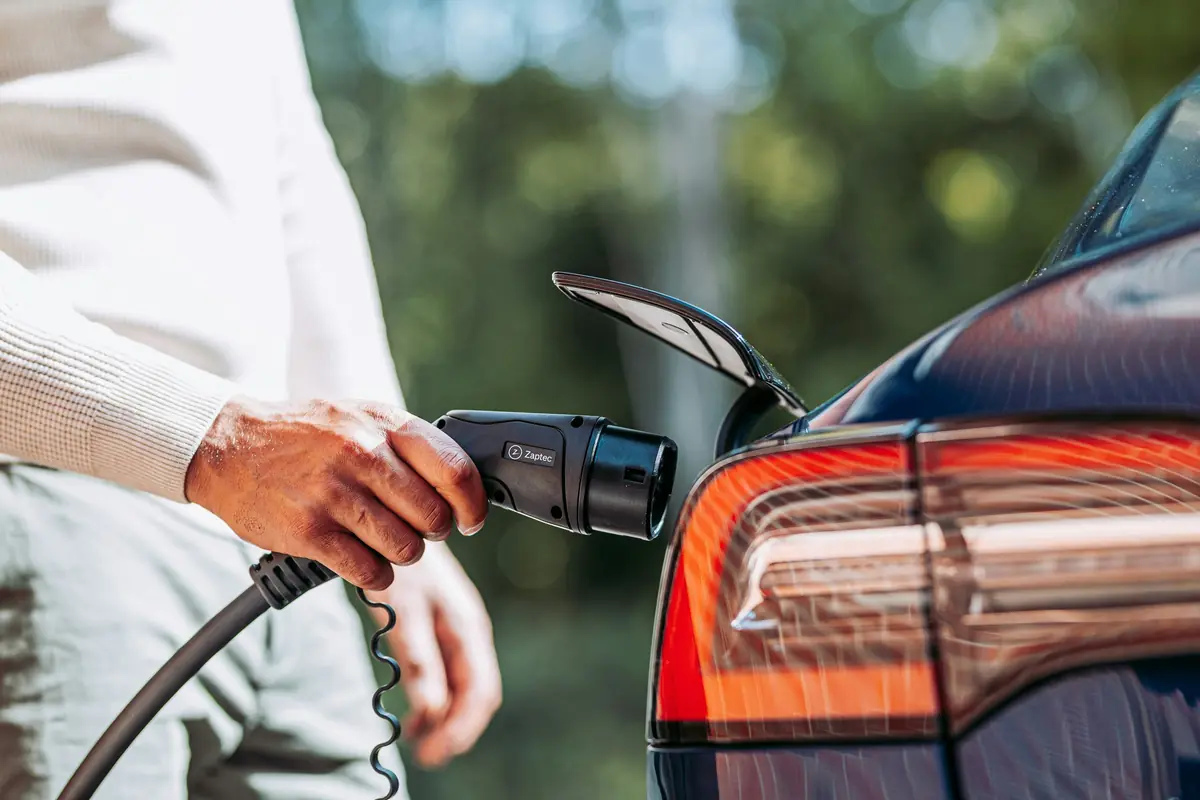 A hand holds a charging connector beside a car
